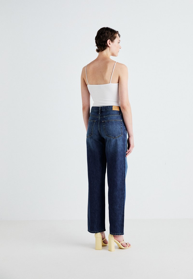 Hochtaillierte, dunkelblaue Jeans aus Denim mit lockerer Passform, kombiniert mit einem weißen Tanktop und gelben Blockabsatzsandalen, von hinten gezeigt.