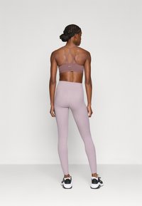 Brassière de sport mauve clair avec bretelles croisées, assortie à un legging taille haute. Des chaussures de sport noir et blanc complètent la tenue.