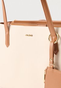 Sac fourre-tout en faux cuir beige avec des poignées marron, des finitions en or et une petite pochette zippée attachée. Logo embossé. Texture lisse.