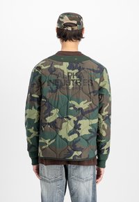 Άτομο που φοράει πράσινη καμουφλάζ καπιτονέ ζακέτα και ταιριαστή καμπύλη, ορατή πλάτη με το κείμενο "Alpha Industries Studio Edition", συνδυασμένο με ανοιχτό τζιν.