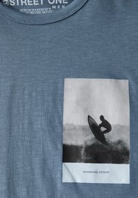 Modré tričko z měkkého materiálu se čtvercovým grafickým vzorem surfaře v černé barvě na bílém pozadí, označené "SUNSHINE ESTATE."