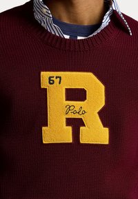 Mörkröd stickad tröja med en stor gul filtpatch föreställande bokstaven "R" och siffrorna "67" samt ordet "Polo" broderat i kursiv.