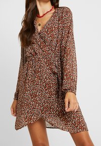 Robe portefeuille marron en tissu léger avec un imprimé animalier dispersé en crème et bleu sarcelle. Manches longues avec poignets froncés et texture douce.