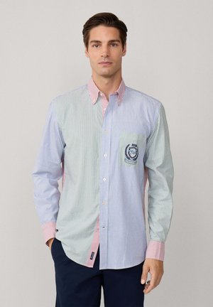 Hombre vistiendo una camisa de rayas multicolores con botones y un emblema bordado en el bolsillo del pecho, combinada con pantalones oscuros, de pie frente a un fondo sencillo.