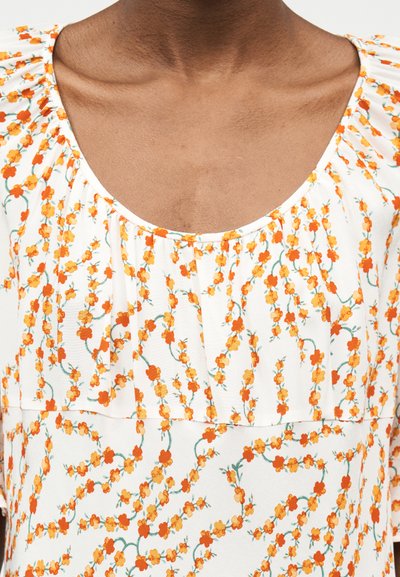 Κοντινή λήψη λευκού μπλουζιού με μικρό πορτοκαλί και κίτρινο λουλουδάτο σχέδιο και στρογγυλή λαιμόκοψη με σούρες σε σκούρο δέρμα.