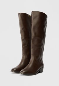 Para ciemno-brązowych, skórzanych butów za kolano na niskim obcasie z bocznymi zamkami, stojących w pozycji pionowej na białym tle.