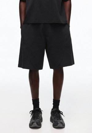 Sorte atletikshorts båret med sorte mellemlange sokker og sorte løbesko på en stående person med hænderne i lommerne.