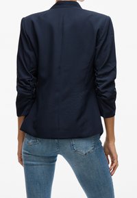 Blazer bleu marine avec une silhouette ajustée, un tissu texturé et un détail froncé sur les manches. Porté avec un jean bleu clair, dévoilant un design épuré au dos.