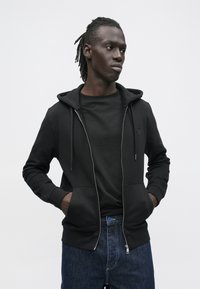 Sudadera negra con cremallera y capucha con cordón, con bolsillos delanteros y textura suave, llevada sobre una camiseta negra y jeans de mezclilla.