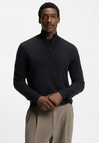 Pull gris foncé à fermeture éclair avec col et poignets côtelés, porté sur un pantalon beige clair. Design simple, texture lisse sans motifs visibles.