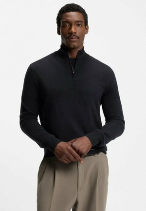 Mörkgrå zip-tröja med ribbad krage och muddar, bärs över ljusbeige byxor. Enkel design, slät textur utan synliga mönster.