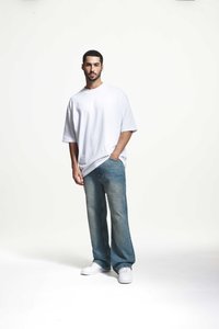 Tricou oversized din bumbac alb, blugi din denim deschis, adidași albi. Modelul stă relaxat cu mâinile în buzunare, pe un fundal simplu.