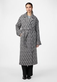 Cappotto lungo a fantasia pied-de-poule in bianco e nero, con colletto ampio, vita con cintura e maniche ampie, abbinato a pantaloni neri.