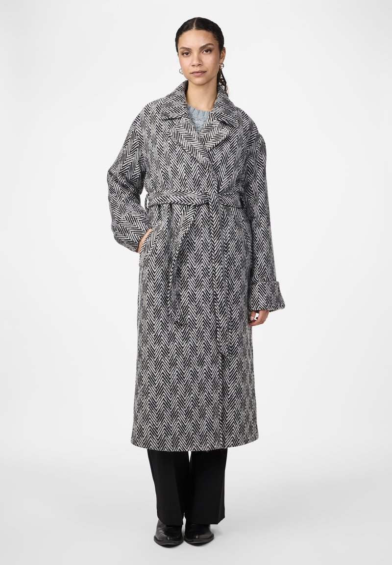 Cappotto lungo a fantasia pied-de-poule in bianco e nero, con colletto ampio, vita con cintura e maniche ampie, abbinato a pantaloni neri.