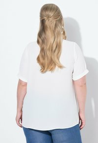 Weiße Bluse mit kurzen Ärmeln aus leichtem Stoff; hochgekrempelte Ärmel; hinten mit abgerundetem Saum; kombiniert mit blauen Jeans; glatte Textur.