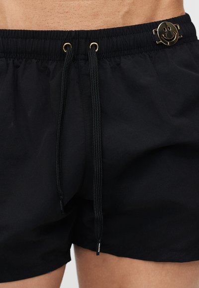 Μαύρα μαγιό σορτς με ελαστική μέση, μαύρα κορδόνια και χρυσό μεταλλικό έμβλημα με χαμογελαστό πρόσωπο κοντά στη μέση στο κάτω μέρος του κορμού ενός ατόμου.