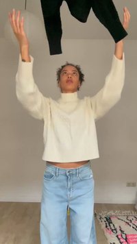 Person trägt einen weißen Pullover und hellblaue Jeans und wirft einen schwarzen Pullover drinnen nach oben, mit einem gemusterten Teppich auf dem Boden.