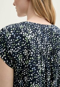 Chemise noire avec un col rond, des manches courtes et un dos plissé. Présente un motif de pois colorés en vert, blanc et bleu.