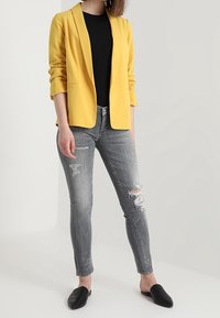 Blazer amarillo con un corte ajustado, mangas largas y solapas en pico, combinado con jeans ajustados grises desgastados y zapatos negros sin cordones.