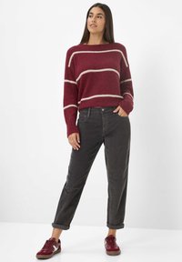 Bordeaux gestreifter Pullover aus weich gestricktem Material, kombiniert mit engen schwarzen Hosen. Das Model trägt bordeauxfarbene Sneakers; lockere Passform mit umgeschlagenen Ärmelenden der Hose.