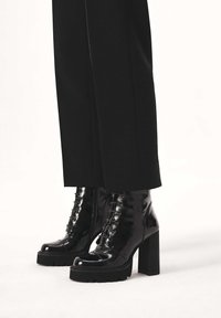 Juodi lako odos kulkšnies batai su stambiais kulnais, priekine raištelių uždarymo dalimi, apvalia nosimi ir briaunuotais padais, derinti su pritaikytais juodais kelnėmis.