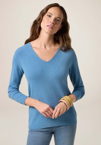 Pull bleu en v en matériau doux, avec des manches longues et des bords côtelés. Assorti à des bracelets multicolores sur un poignet.
