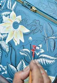 Blauwe leren tas met florale ontwerpen, met geschilderde gele bloemen, groene bladeren en een vlinder. Penseel-detail dat vakmanschap benadrukt.