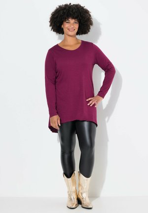 Tunique bordeaux à manches longues avec un ourlet arrondi, associée à des leggings noirs brillants et des bottes de cowboy dorées, se tenant devant un fond blanc.
