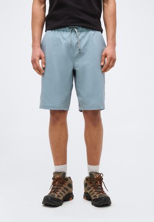 Isik, kes kannab helerohelisi nööriga lühikesi pükse, musta t-särki, valgeid sokke ja pruune matkasaapaid, seisab ühevärvilise tausta ees.