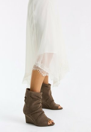 Jambes dans une jupe blanche bordée de dentelle, portant des bottines compensées taupe en daim à bout ouvert sur fond blanc.