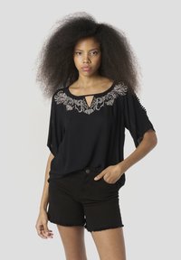 Top negro de manga corta con escote en forma de ojo de cerradura, detallado con bordado floral blanco en el pecho, combinado con pantalones cortos de mezclilla negros.
