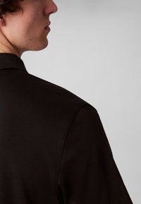 Crna polo majica sa strukturiranim ovratnikom, glatkom teksturom i suptilnim šavovima, gledana s boka, ističući izrez i rame.