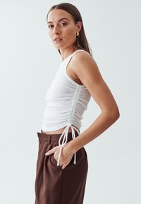 White ribbed tank top with side ruching and adjustable ties, paired with brown trousers. Smooth texture and fitted design.