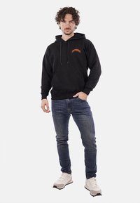 Felpa nera con tasca frontale, cappuccio con cordino e logo arancione. Indossata con jeans in denim blu e scarpe da ginnastica bianche.