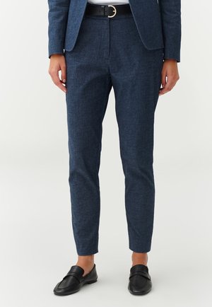 Femme portant un pantalon bleu marine sur mesure, une ceinture noire avec une boucle dorée, des mocassins noirs et un blazer bleu marine assorti.