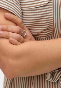 Anello in metallo argento con design curvo e organico, caratterizzato da un piccolo accento a forma di perla. Indossato su una mano su uno sfondo di tessuto a righe.