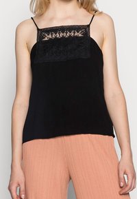 Camisola nera con delicata sovrapposizione di pizzo sullo scollo, spalline sottili e una vestibilità rilassata. Indossata con pantaloni chiari a coste.