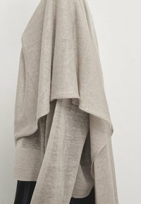 Jasno beżowy sweter z dzianiny o luźnym kroju, z wyraźną fakturą tkaniny i warstwową konstrukcją. Posiada szerokie rękawy i żeberkowany dół.