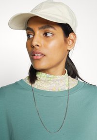 boné beige claro, sweatshirt verde, turtleneck floral em camadas, colar de corrente prateada, textura suave, design casual.