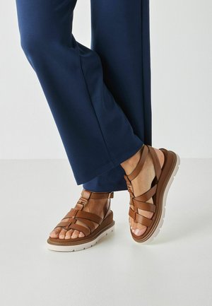 Brązowe sandały na paski z grubą białą podeszwą i ozdobnymi klamrami. Cholewka z fakturowanej skóry z otwartym noskiem, zestawiona z granatowymi spodniami.