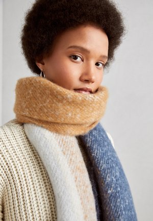 Jeune femme aux cheveux courts et bouclés portant des boucles d'oreilles en anneau et une grosse écharpe multicolore par-dessus un pull en maille crème, sur fond uni.