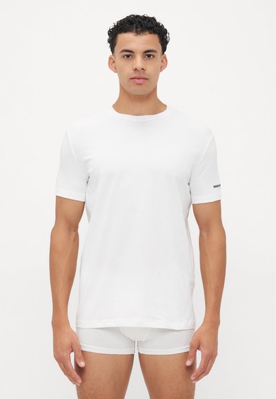 Weißes T-Shirt mit kurzen Ärmeln aus glattem Baumwollstoff, mit Rundhalsausschnitt und einem kleinen schwarzen Logo auf dem linken Ärmeln. Kombination mit weißen Shorts.