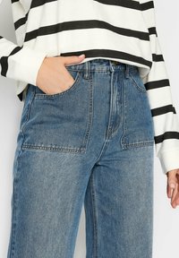 Jeans de mezclilla azul de alta cintura con un acabado deslavado, que presentan grandes bolsillos delanteros de parche y sutiles detalles de costura. Llevados con una camiseta a rayas.