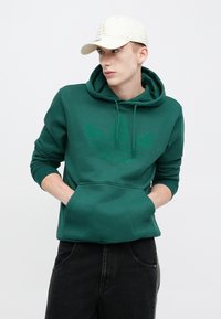 Πράσινο φούτερ με κουκούλα, με υφασμένο σκούρο πράσινο λογότυπο στο εμπρός μέρος, τσέπη τύπου καγκουρό και ρυθμιζόμενο κορδόνι στην κουκούλα. Φορεμένο με μαύρο τζιν.