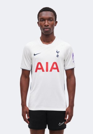 Homme portant un maillot de football blanc Tottenham Hotspur avec un logo rouge AIA et un short noir Nike, debout, les bras détendus, sur un fond uni.