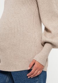 Beige ribbestrikket genser med lange ermer og tettsittende mansjetter, båret over blå denimjeans som fremhever den myke teksturen og den figurnære designen.