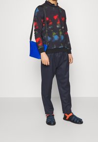 Sudadera de tie-dye multicolor con acentos en negro, combinada con pantalones tapered en azul marino y bolso cruzado azul. Sandalias negras con tiras naranjas.
