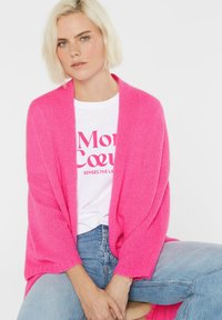 Blonde Frau, die eine leuchtend pinke Strickjacke über einem weißen T-Shirt mit Grafik und hellblauen Jeans trägt, sitzt auf einem Holzschemel.