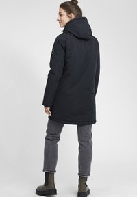 Abrigo largo negro con capucha y textura suave, con una cintura ajustada, bolsillos laterales y un logo en el brazo izquierdo. Jeans grises y botas de tobillo.