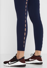 Marina leggings med utskärningar längs sidan, kombinerade med svarta och röda träningsskor med vita detaljer och en grå sula.